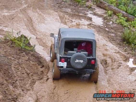Black Jeep Wrangler with a soft top driving through mud, rear view. Spare tire visible