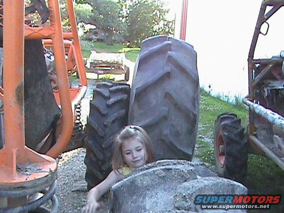 Girl peeking over a large tire with orange and red off-road vehicles in the background