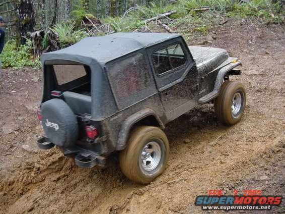 Muddy 1995 Jeep Wrangler with a soft top, viewed from the rear three-quarter angle on a dirt trail