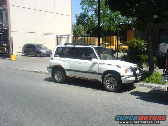 White SUV with rust on the lower body panels parked on a street. Another gray minivan is visible in the background