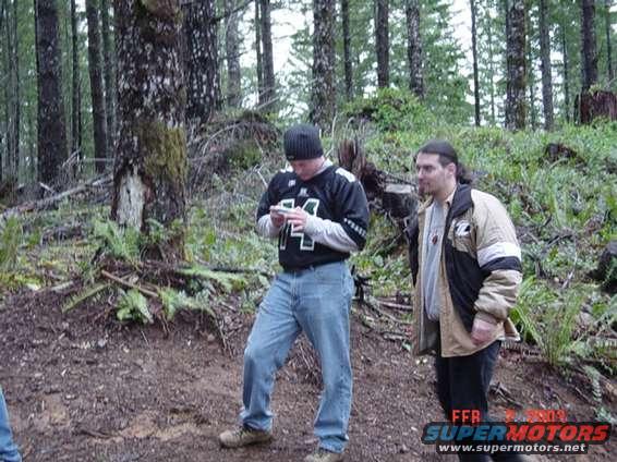 Two guys stand in a wooded area, one looking at his phone