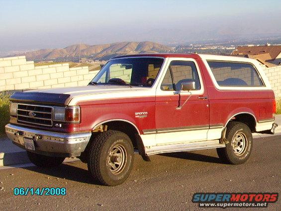 1990 Ford Bronco parked on asphalt with a brick wall and hills in background