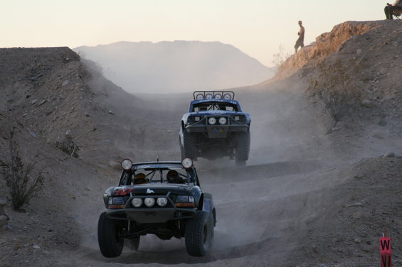 Two off-road trucks race up a dusty hill, one behind the other