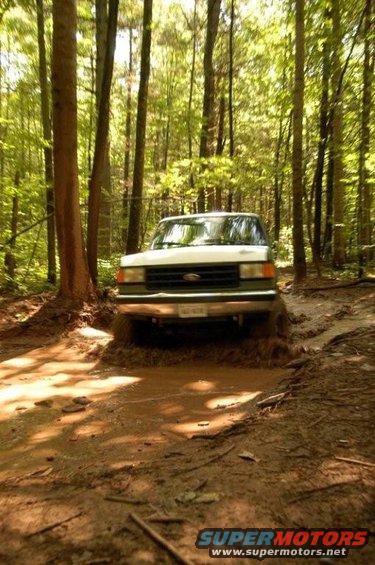 1987 Ford Bronco driving through a muddy trail in a forest