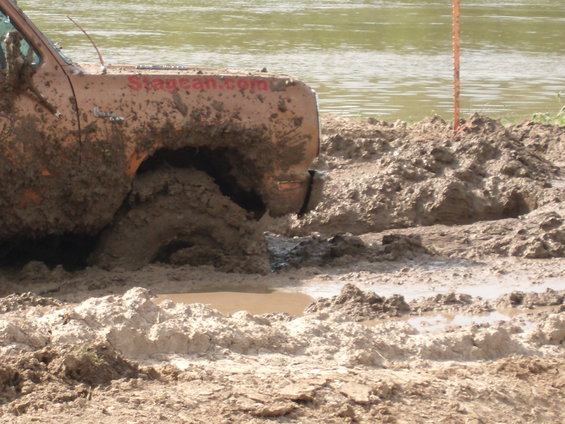 Orange truck stuck in mud, front wheel deep in mire, with "Stagecan.com" visible on the door