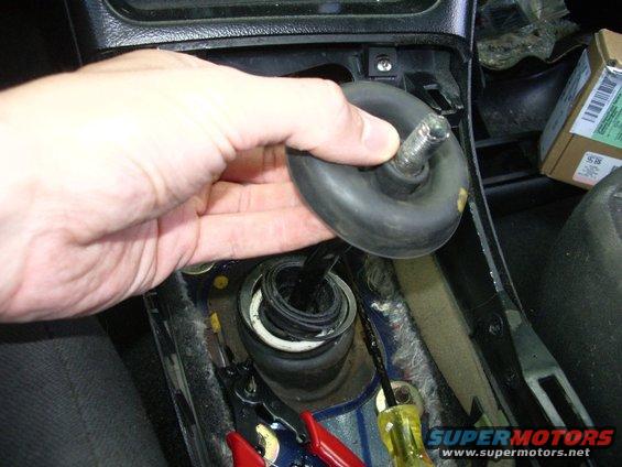 Hand holding black rubber boot over car's shifter opening, tools visible below