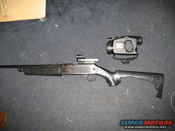 Black shotgun with a scope and a night vision device, lying on a dark carpet. The stock is wrapped in black tape