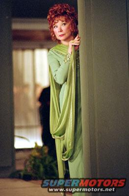 Woman with red curly hair in a green dress peeks from behind a green curtain