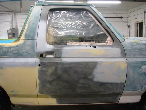 Side view of a 1992 Ford Bronco in a body shop, covered in primer and masking plastic