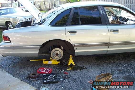 Silver sedan, hood and trunk open, rear wheel off, jack and tools on ground. Another car is parked behind it