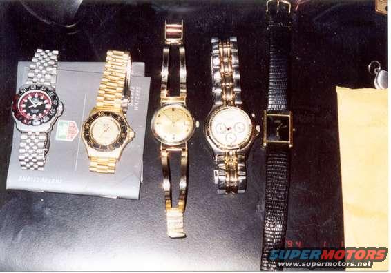 Five watches on a dark surface, including a silver watch with a red bezel and a gold watch with a black bezel