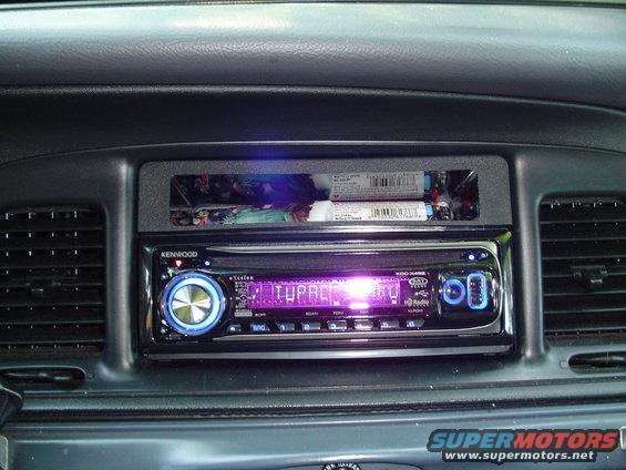 Car dashboard with a Kenwood stereo displaying 'TUPAC' and a storage compartment above it