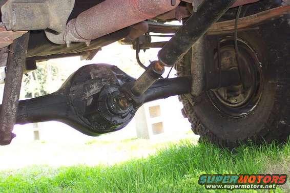Underside view of a vehicle's rear axle, differential, driveshaft, and tire, with grass in the foreground