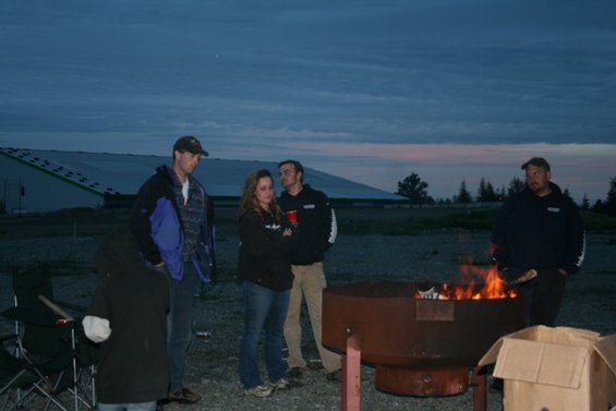 People stand around a large fire pit at dusk, with a building in the background