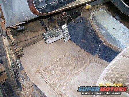 Vehicle interior showing brake and accelerator pedals, floor mat, and part of the driver's seat