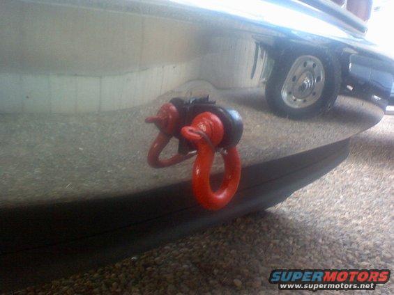 Red tow hook mounted on a vehicle's front bumper. A wheel is visible in the background