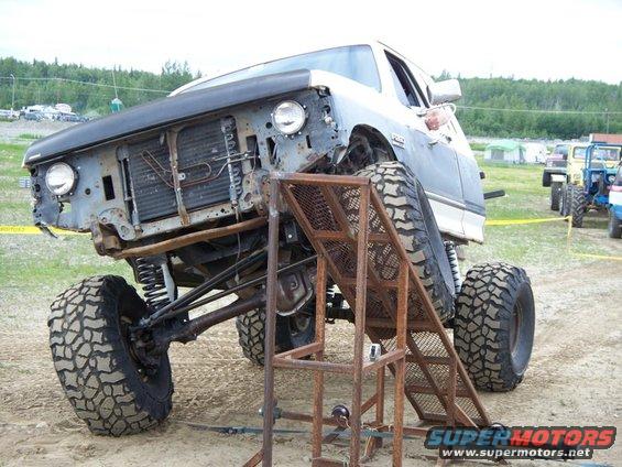 Modified pickup truck with large tires on a metal ramp. Other vehicles in background
