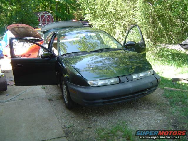 1997 Saturn SC with driver's door and passenger door open, parked outside. Another car is visible in the background with its hood up