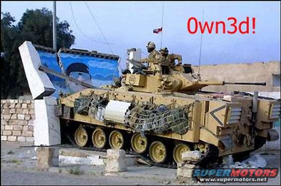 Tan military tank with netting and a cannon, angled from the side, with a broken blue wall behind it
