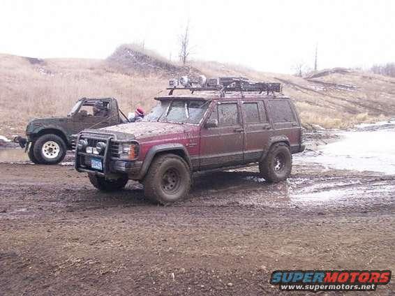 Maroon Jeep Cherokee SUV with roof rack and brush guard, parked in mud