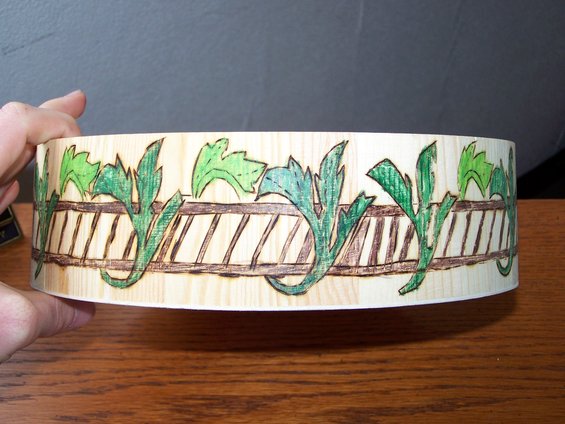 Wooden cylinder with a decorative border of green leaves and brown fence-like pattern, held by a hand