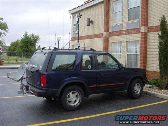 Dark blue SUV with a satellite dish mounted on the rear bumper, parked in a lot next to a brick building