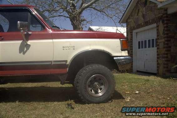 Side view of a two-tone red and white Ford Bronco parked next to a brick garage