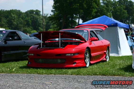Red sports car with hood and trunk open, parked on grass next to a black car and a tent