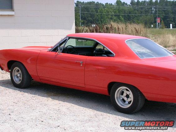 Red 1970 Plymouth Barracuda coupe parked on gravel next to a white building