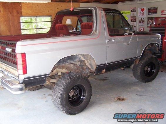 Silver Ford Bronco with lifted suspension and large tires, viewed from the rear three-quarter angle inside a garage