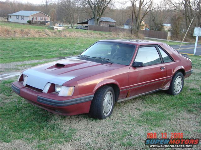 Red coupe with a hood scoop parked on grass. The front bumper is gray