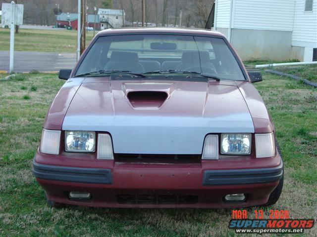 Front view of a red and grey 1984 Ford Mustang with a hood scoop and aftermarket headlights