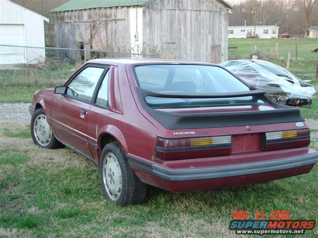 Maroon Ford Mustang parked on grass with a second car covered in plastic behind it. Buildings and trees in background