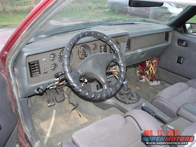 1984 Ford Mustang interior showing dashboard, steering wheel, seats, and exposed wiring under the dash