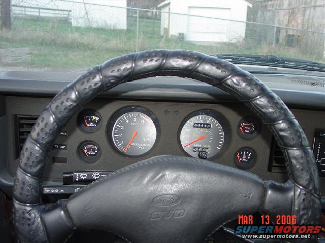 Car dashboard with a leather-wrapped steering wheel, tachometer, speedometer, and various gauges. A watermark reads 'MAR 13 2006 SUPERMOTORS'
