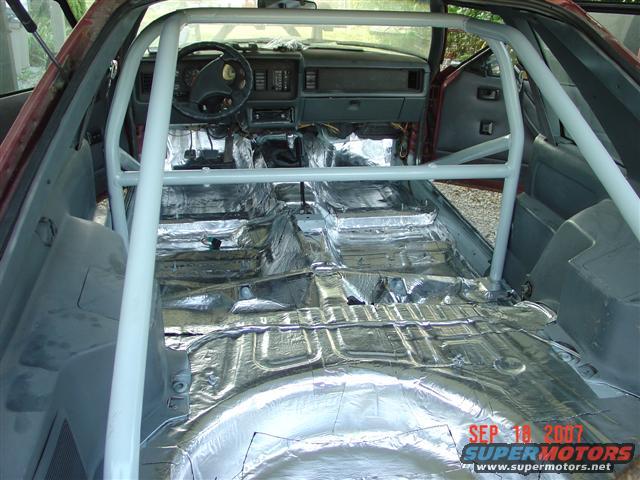 1984 Ford Mustang interior with roll cage, sound deadening on floor, gray dashboard, red exterior visible