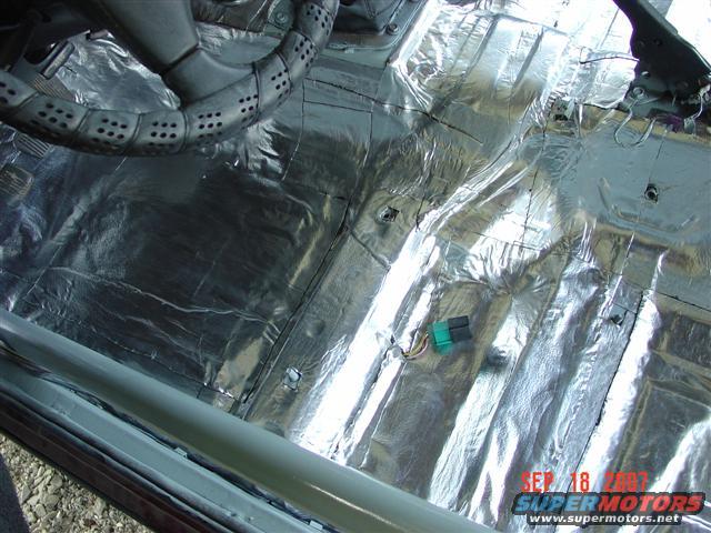 Car interior floor covered in silver sound deadening material, with part of the steering wheel visible
