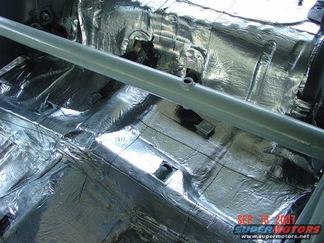 car interior floor covered in silver sound deadening material with a metal bar across the middle