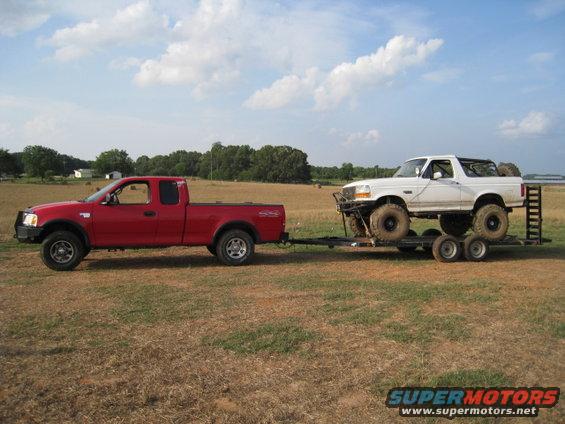 Red Ford F-150 pickup towing a white 1996 Ford Bronco on a trailer. Both vehicles are off-road modified