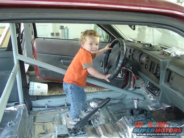 Interior of a 1984 Ford Mustang with a child at the steering wheel and roll cage installed
