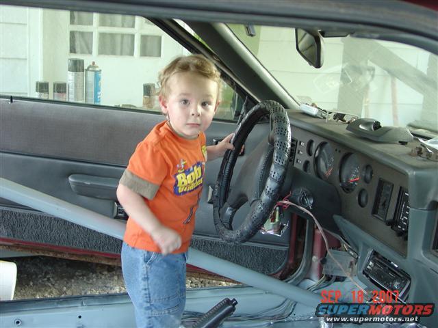 Child standing in the interior of a 1984 Ford Mustang, holding the steering wheel