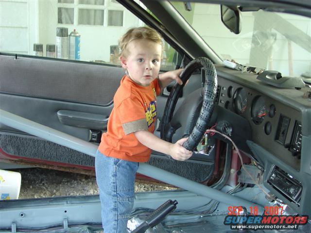 Child holding steering wheel inside a 1984 Ford Mustang interior. Roll cage visible