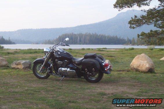 Black motorcycle with saddlebags parked on grass near a lake and mountains