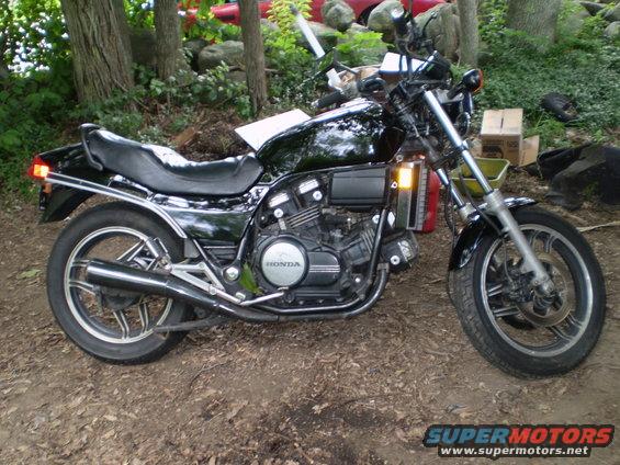 Black Honda motorcycle parked on dirt, trees and debris in background. Supermotors.net logo visible