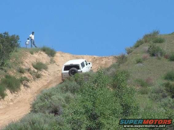 White SUV with spare tire on back driving down a dirt hill, two people stand on the ridge above