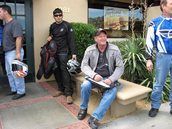 Motorcycle riders with helmets and gear stand and sit outside a building