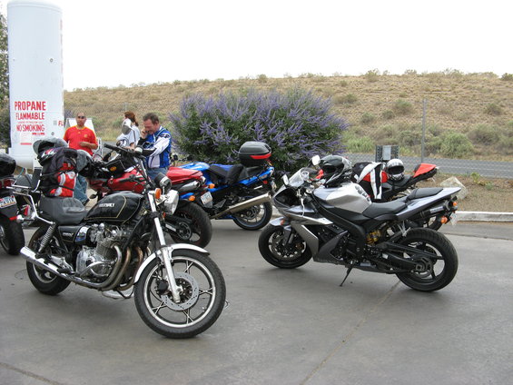 Several motorcycles are parked in front of a large propane tank, with people standing around and a flowering bush in the background