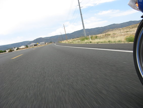 Close-up of a blue motorcycle wheel and tire on a paved road, with mountains and houses in the background