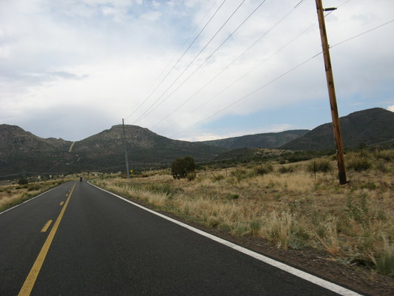 Two-lane asphalt road with yellow and white lines, passing through a dry, grassy landscape with hills and power lines under a cloudy sky