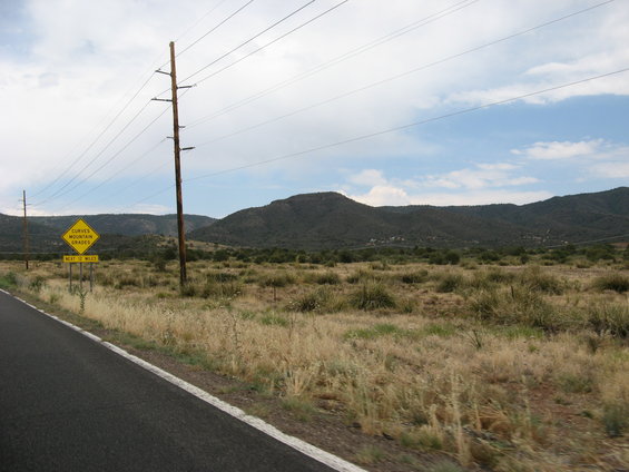 Roadside view with dry grass, power lines, and distant mountains under a cloudy sky. A yellow sign warns of curves and mountain grades